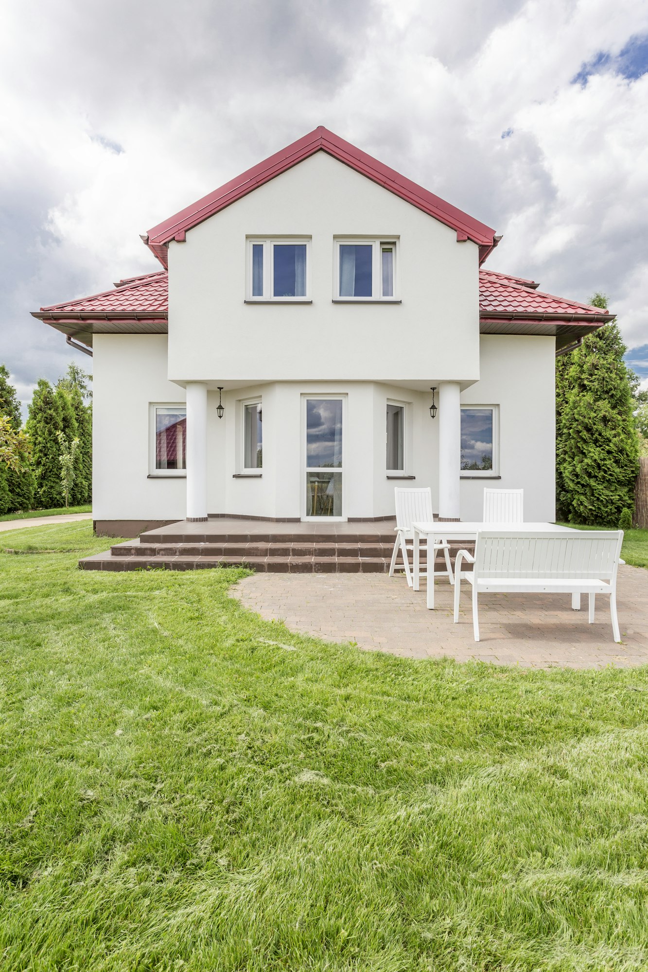 Modern two-story house with a red roof, white walls, and large windows, featuring a terrace with white outdoor furniture on a grassy lawn.