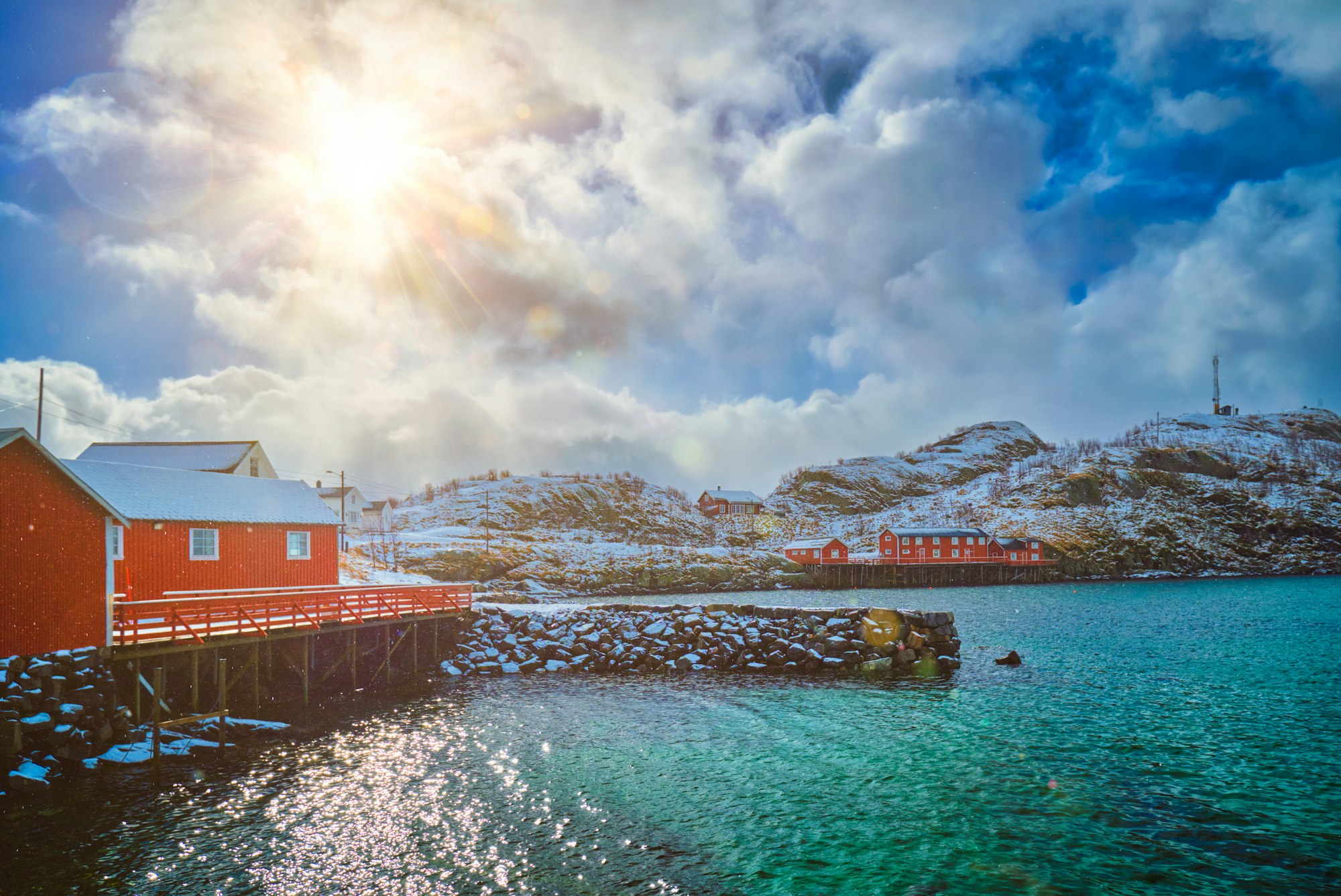 Red rorbu houses on stilts by the water in Lofoten Islands, Norway, with snow-covered hills and a bright sun illuminating the scene.